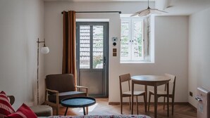 Ein Zimmer mit Bett, Sessel, kleinem Tisch, Esstisch, Deckenventilator und einem Fenster mit Vorhängen.