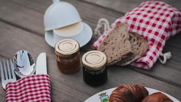 A wooden table with a white cup, two jars, a bread roll, a croissant, and a folded napkin.