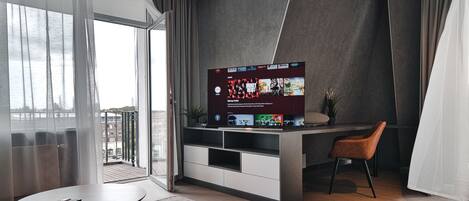 A modern living room with a large flat-screen TV mounted on a wall, a small round table, a brown leather chair, and a view of the city through large windows.