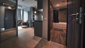 Habitación de hotel moderna con piso de madera, un espejo grande, una puerta de madera oscura y un baño visible.