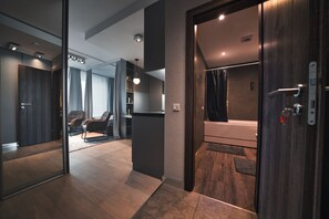 A modern hotel room with a wooden floor, a large mirror, a dark wooden door, and a visible bathroom.