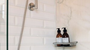 Casa de banho com porta de vidro, paredes revestidas a azulejo branco, parede de detalhes em mármore, chuveiro com mangueira e duas garrafas numa prateleira.