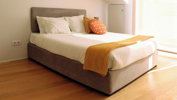 Cama com cabeceira, travesseiros e cobertor num quarto com piso de madeira e janela.