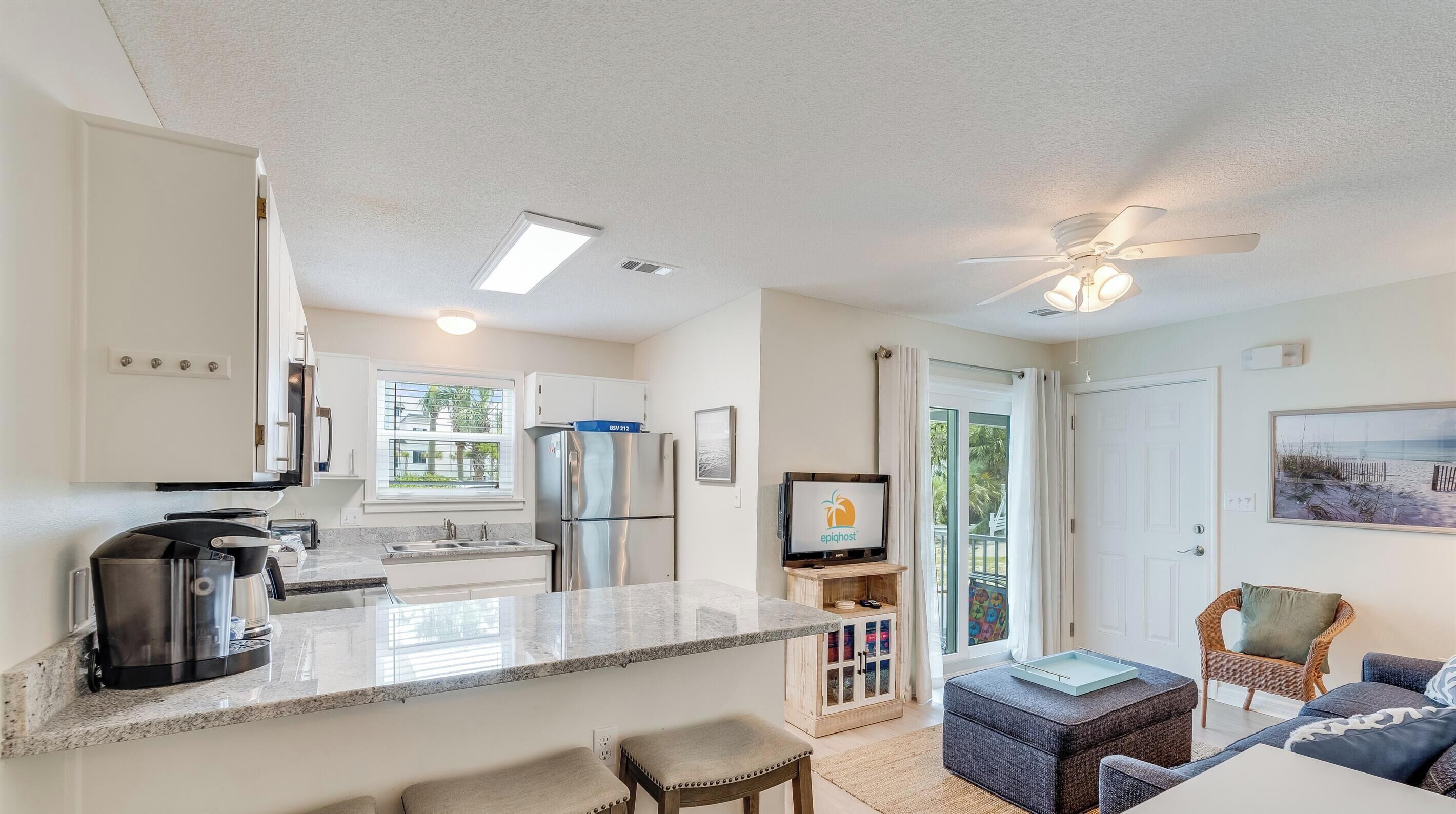 A modern kitchen with a central island, bar stools, a dining area with a white table, and a living room with a sofa and a wicker chair.
