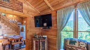 Interior de una cabaña de madera con comedor, televisor montado en la pared y vistas al exterior.