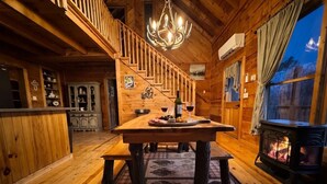 Interior de una cabaña de madera con mesa de comedor, banco, candelabro, chimenea y decoración rústica.