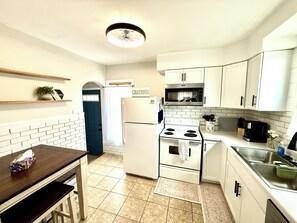 A compact kitchen with white cabinets, a white refrigerator, a microwave, and a stove. There is a small dining table with a chair, a blue door, and a ceiling fan. - Blue Bungalow (Cedar Rapids)