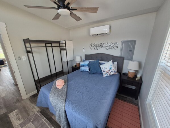 A bedroom with a bed, a ceiling fan, an air conditioning unit, a nightstand, a lamp, and a decorative wall piece.