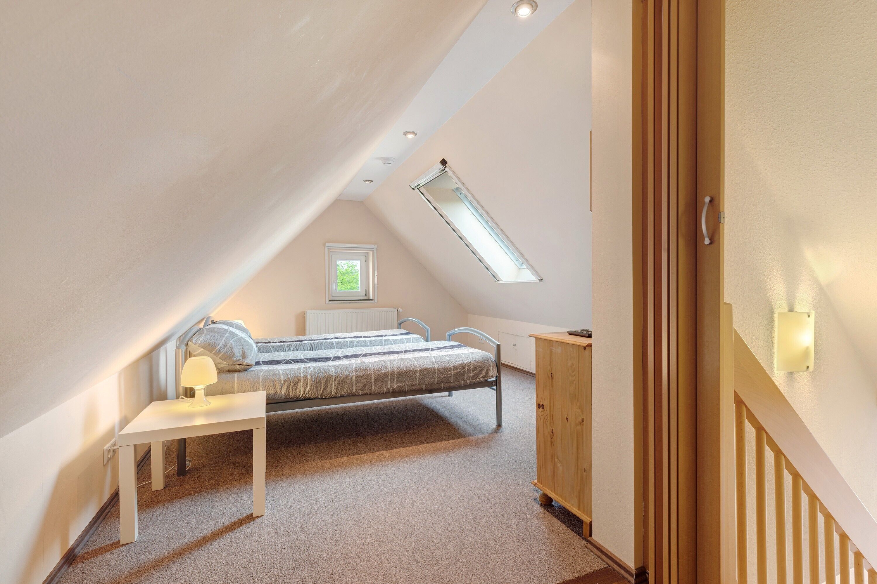 Une chambre avec un plafond en pente, un lit, une table de chevet avec une lampe, une fenêtre et une armoire en bois.