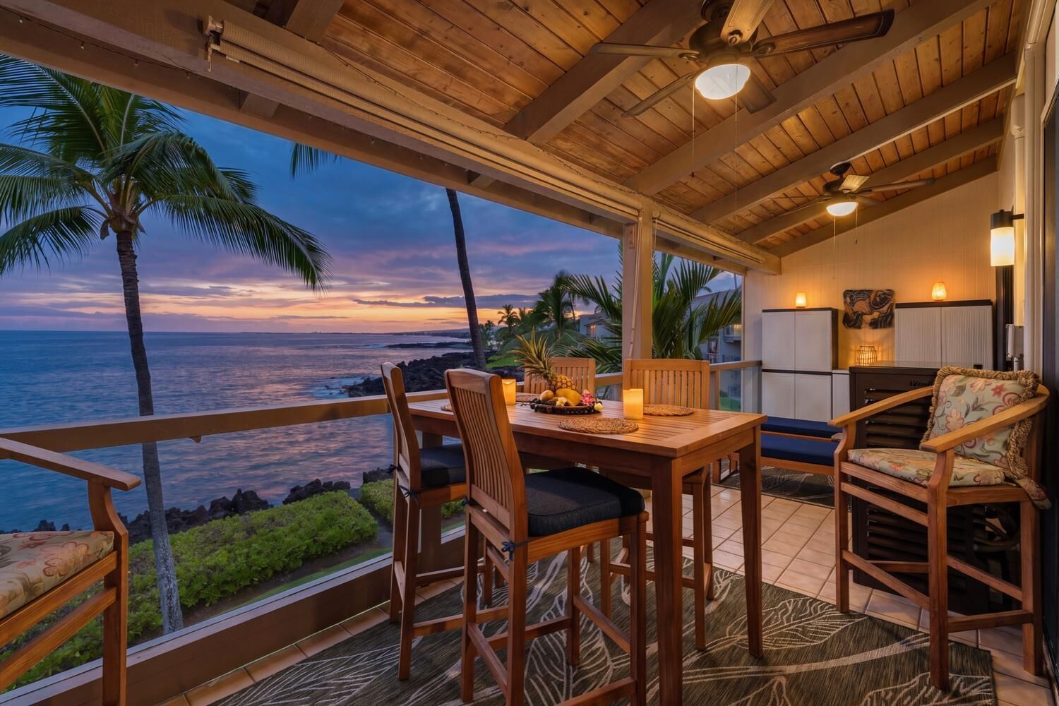 A wooden deck with a table and chairs, overlooking a coastal view with palm trees and the ocean.