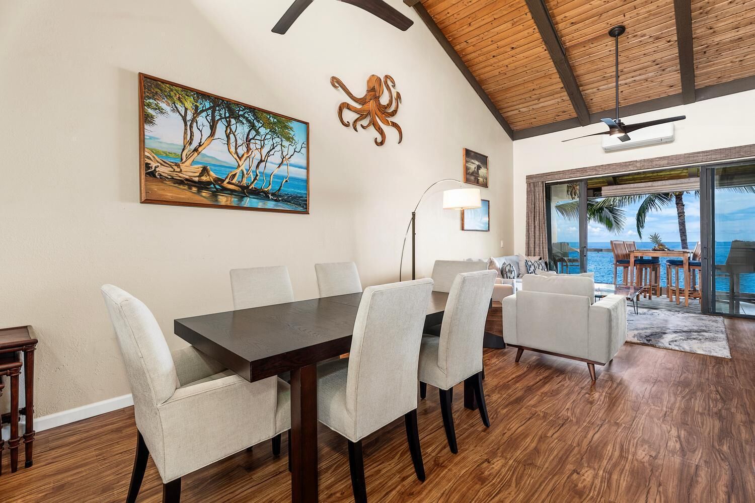 A dining area with a wooden table and chairs, a painting of a tree by the sea, and a view of the ocean through large windows.