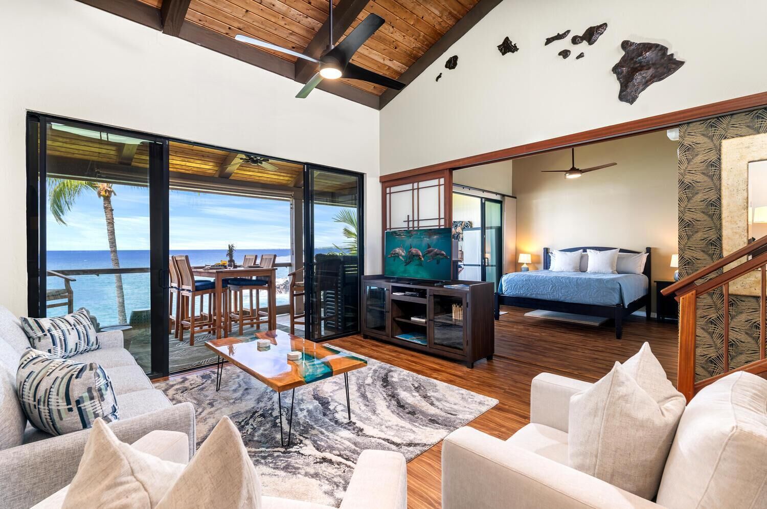 A spacious living area with a large TV, a balcony with ocean views, a bedroom with a bed and nightstand, and a staircase.