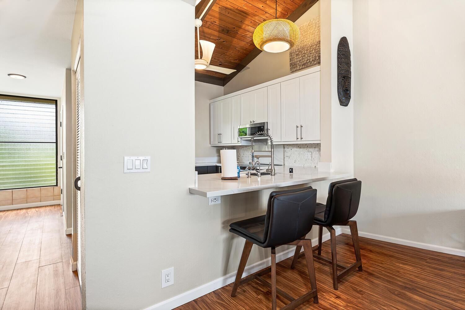A compact kitchen with white cabinets, a microwave, and a sink. There are two black chairs and a wooden ceiling.
