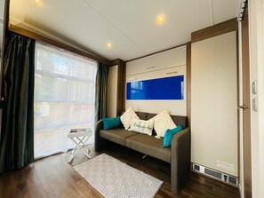 A room with a sofa, a small table, a patterned rug, a large window with curtains, and a wall-mounted TV. - Ravenswood Pod near Fort William (Fort William)