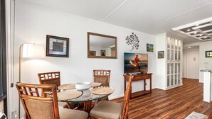 A dining area with wicker chairs, a glass table, and a television on a wooden cabinet. - Kona Pacific E629: Beautifully Remodeled Condo! (Kailua-Kona)