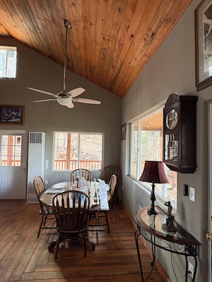 A dining area with a wooden ceiling, a ceiling fan, a dining table with chairs, a clock, and windows with a view of trees. - Terrific 2-bedroom country house in charming Woodland Park (Woodland Park)