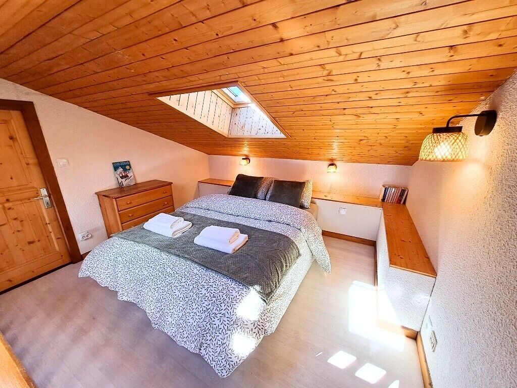 Une chambre avec un plafond en pente, un puits de lumière, un lit recouvert d’une couvre-lit à motifs, des tables de chevet et des lampes fixées au mur.