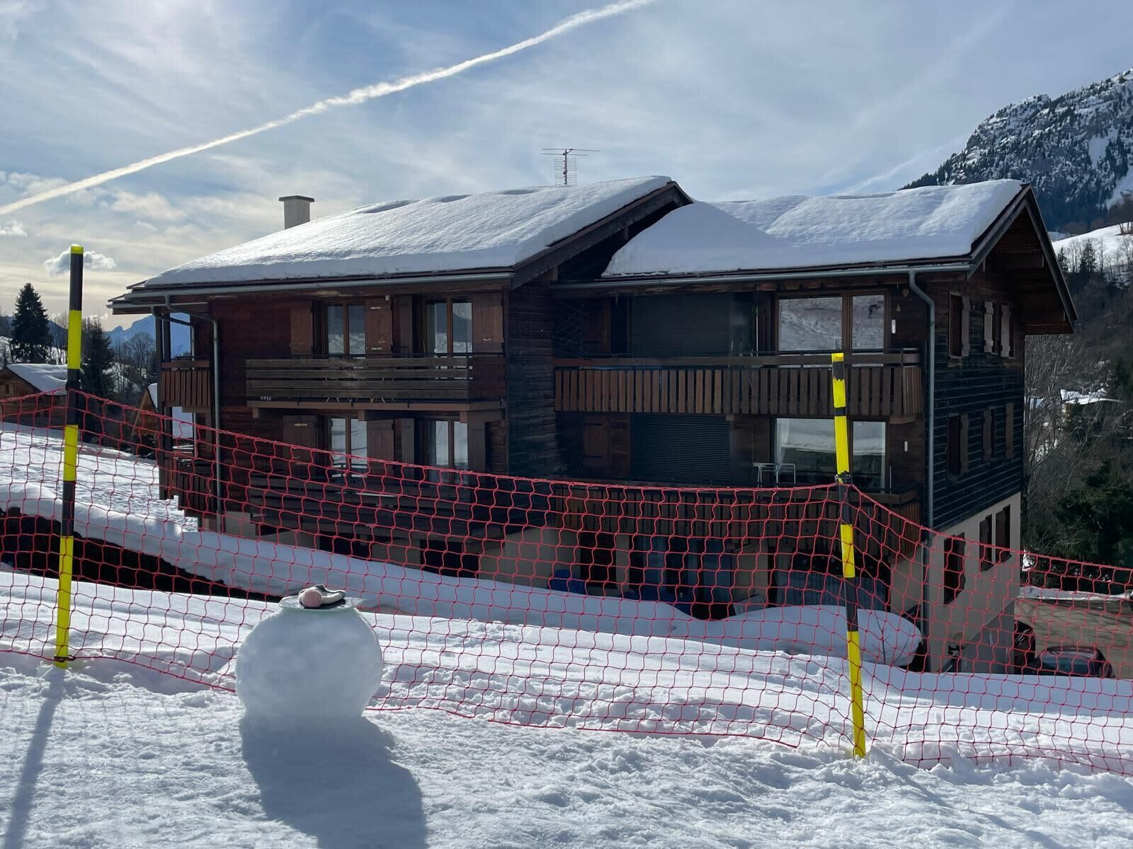 A wooden chalet with a balcony, surrounded by snow, and a red net fence in the foreground.