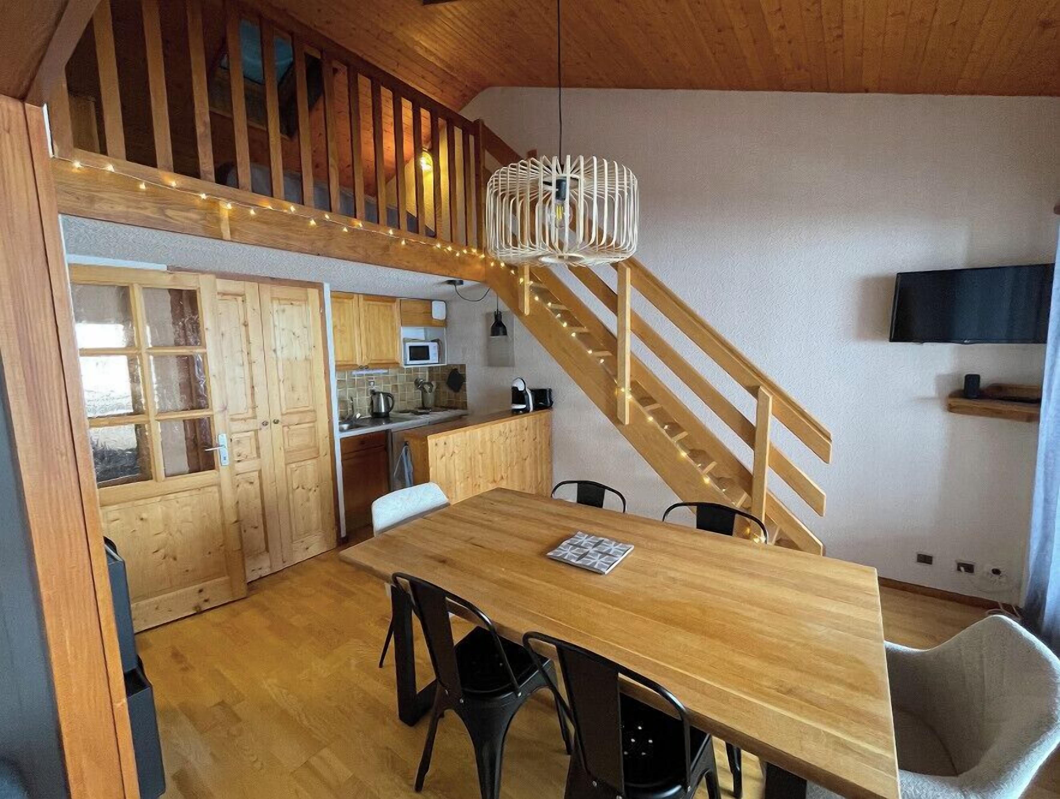 Une table à manger en bois avec des chaises, un coin cuisine avec des meubles en bois, un escalier et un téléviseur fixé au mur.