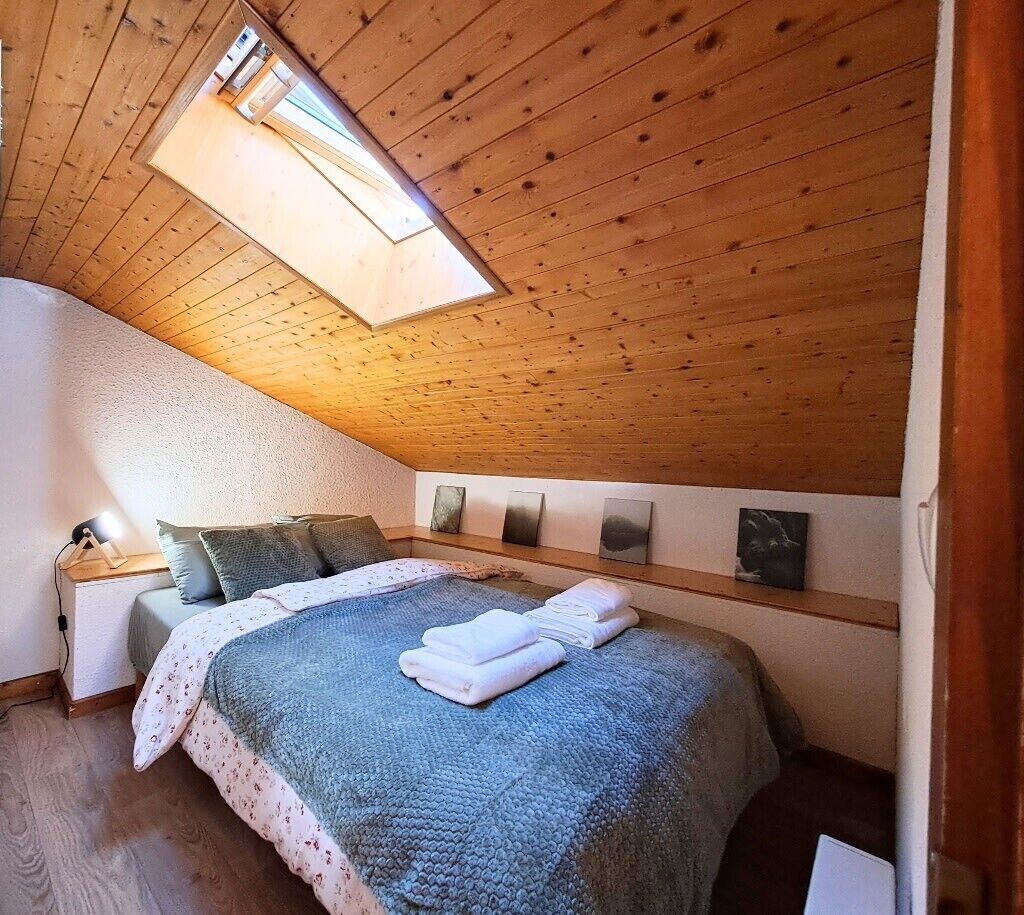 Een slaapkamer met een schuine kap, een dakraam, een bed met een blauwe deken, een nachtkastje en een kunstwerk aan de muur.