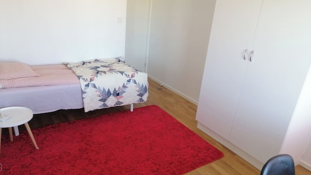 A room with a bed, a red rug, a small white table, and a wardrobe. - Room-cozy Guest Room in Green Uppsala - (Uppsala)