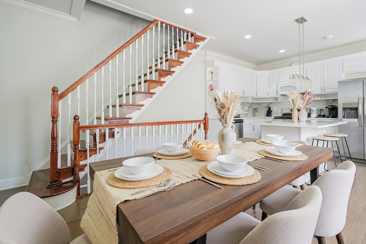 A dining area with a wooden staircase, white cabinetry, and a kitchen in the background.