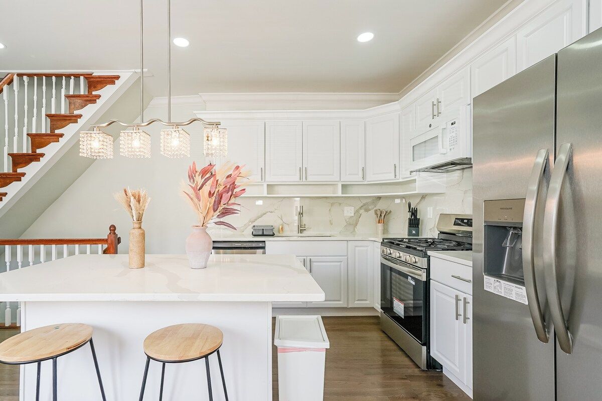 A modern kitchen with white cabinetry, a stainless steel refrigerator, a central island with stools, and a staircase with wooden railings.