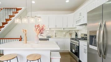 A modern kitchen with white cabinetry, a stainless steel refrigerator, a central island with stools, and a staircase with wooden railings.