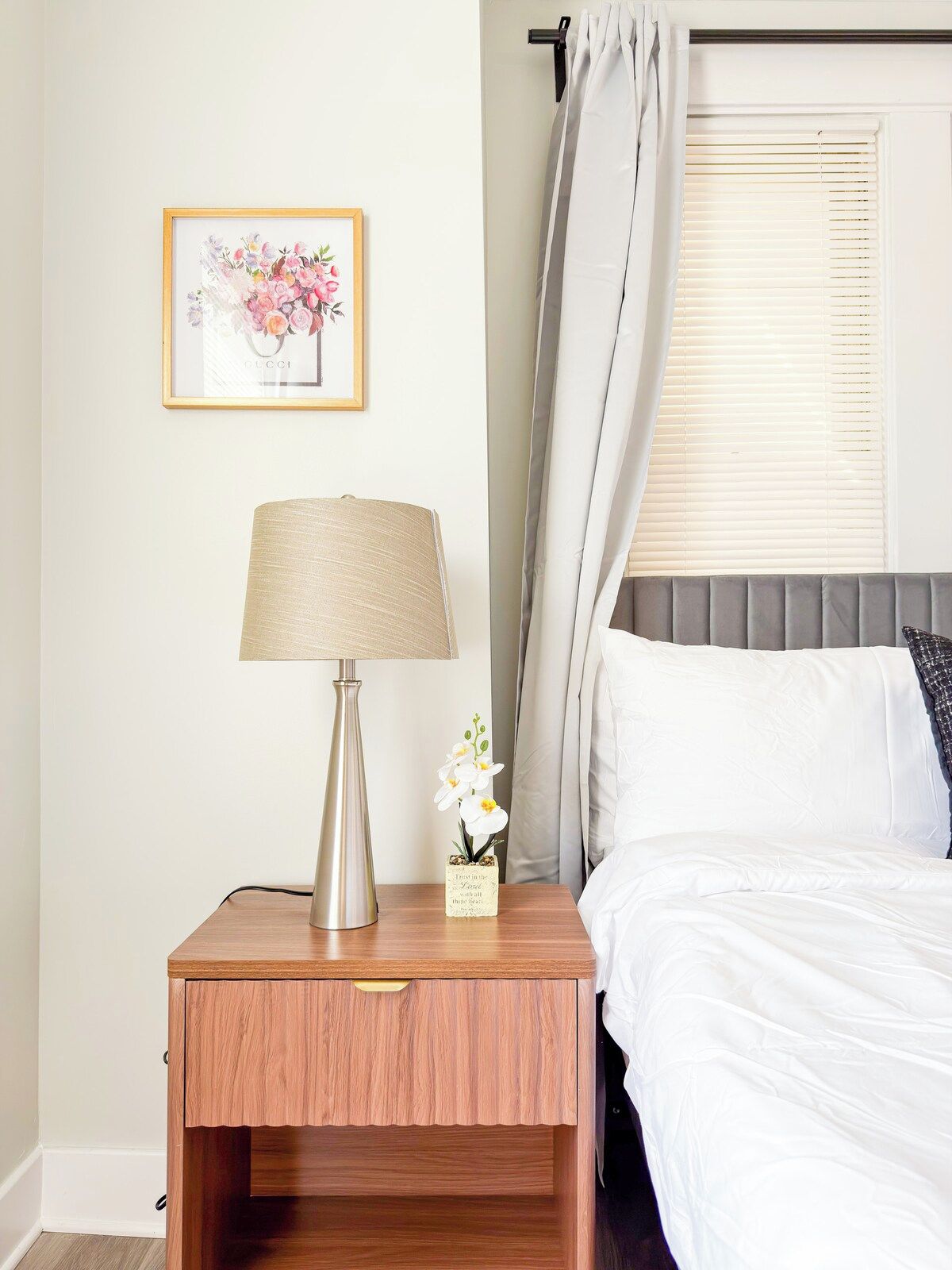 A neatly made bed with white linens, a bedside lamp, a framed picture on the wall, and a window with blinds.