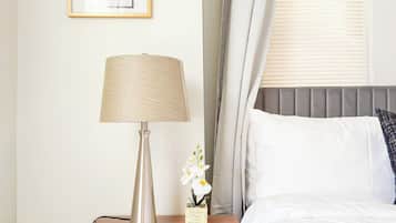 A neatly made bed with white linens, a bedside lamp, a framed picture on the wall, and a window with blinds.