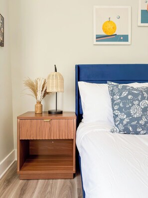 A bedroom with a wooden nightstand, a lamp, a vase with dried plants, a bed with a blue headboard, and a floral pillow.