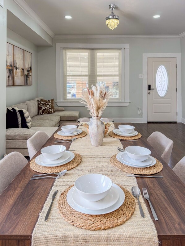 A dining room with a wooden table set for six, surrounded by chairs, with a vase of dried flowers in the center.