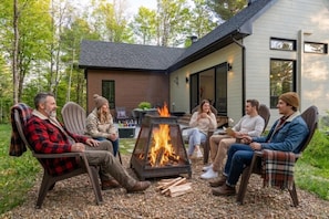 A group of people seated around a fire pit in an outdoor setting with a house in the background. - Le Pin | Private Spa & Billiards | Nature & Golf (Brownsburg)