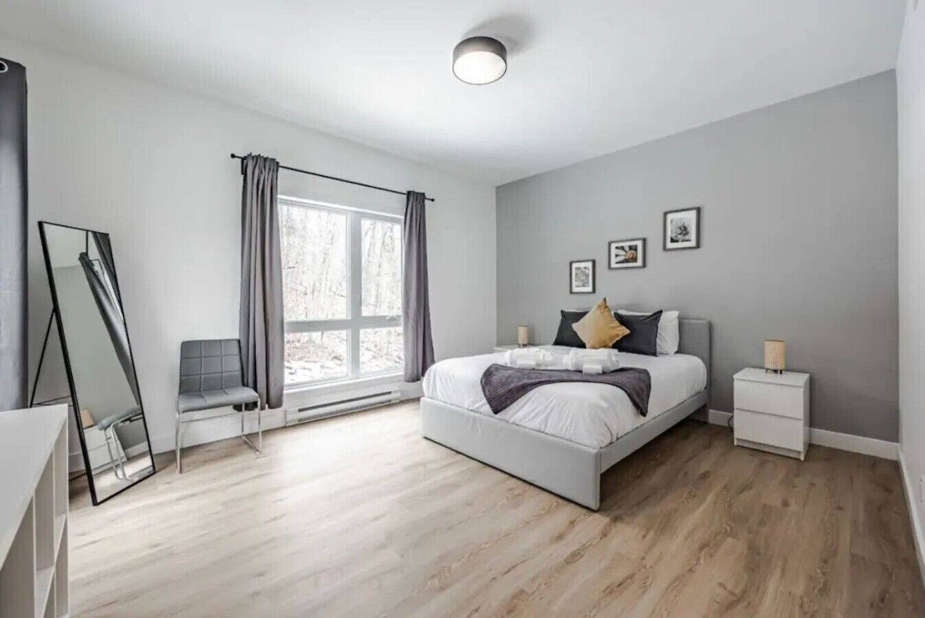 A modern bedroom with a large bed, a chair, a bedside table, a nightstand, a mirror, and a window with curtains.