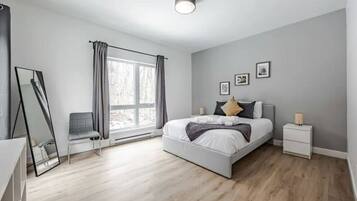 A modern bedroom with a large bed, a chair, a bedside table, a nightstand, a mirror, and a window with curtains.
