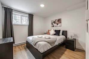 A modern bedroom with a large bed, a bedside table, a nightstand, a window with curtains, and a wall-mounted artwork. - Le Pin | Private Spa & Billiards | Nature & Golf (Brownsburg)