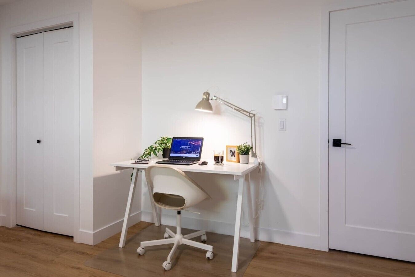 A small, white desk with a laptop, a desk lamp, and a chair in a room with wooden flooring and white walls.
