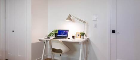 A small, white desk with a laptop, a desk lamp, and a chair in a room with wooden flooring and white walls.