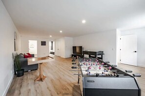 A modern living room with a foosball table, a grey sofa, and a flat-screen TV. - Le Pin | Private Spa & Billiards | Nature & Golf (Brownsburg)