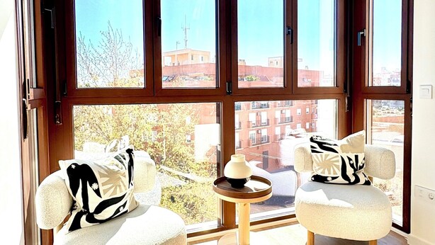 A room with large windows, two armchairs, a small round table, and a view of buildings outside. - New Modern Retreat at Malvarrosa Beach (València)