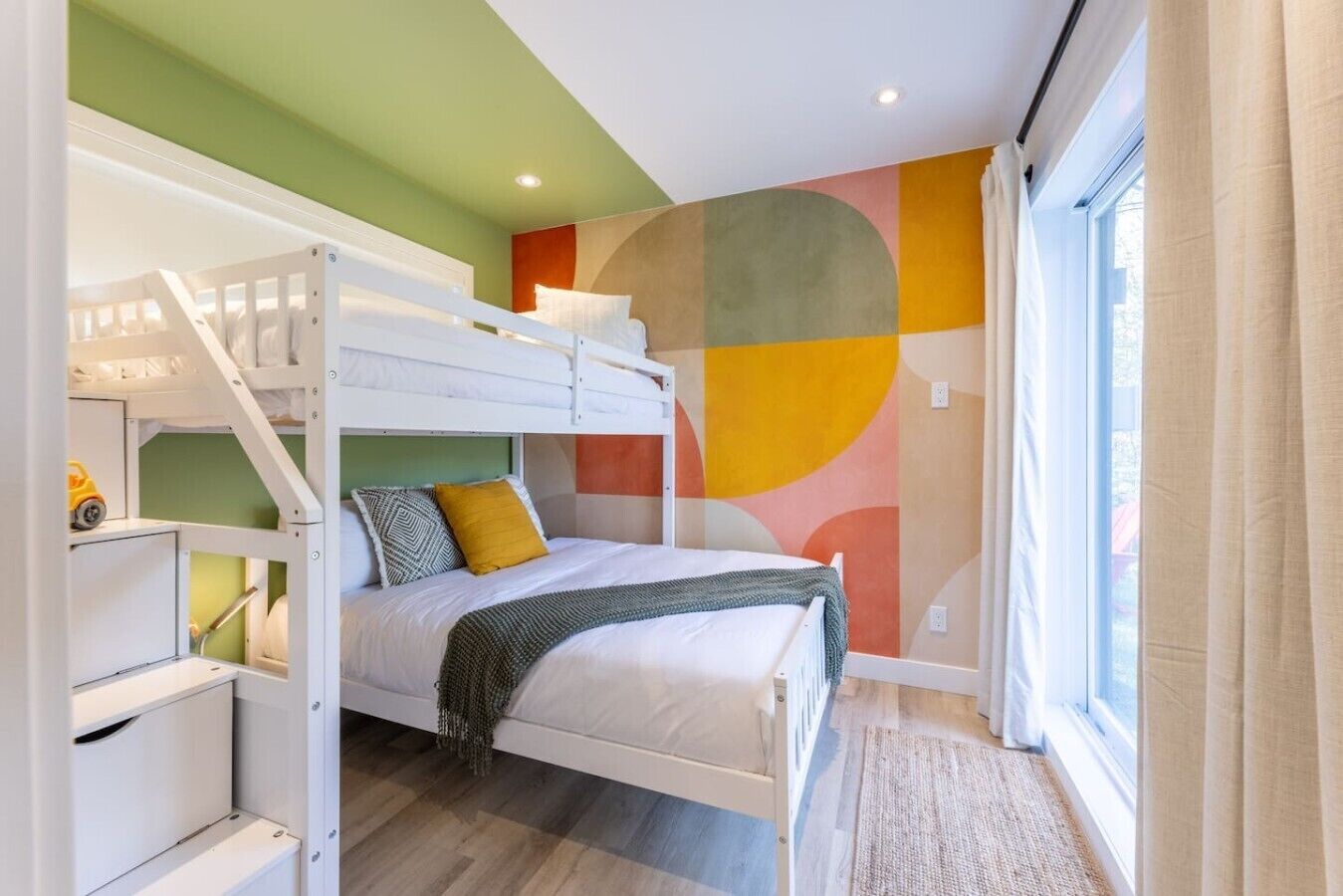 Une chambre avec des lits superposés, un mur décoré d’une fresque abstraite aux couleurs vives, une fenêtre avec des rideaux et un plancher en bois.