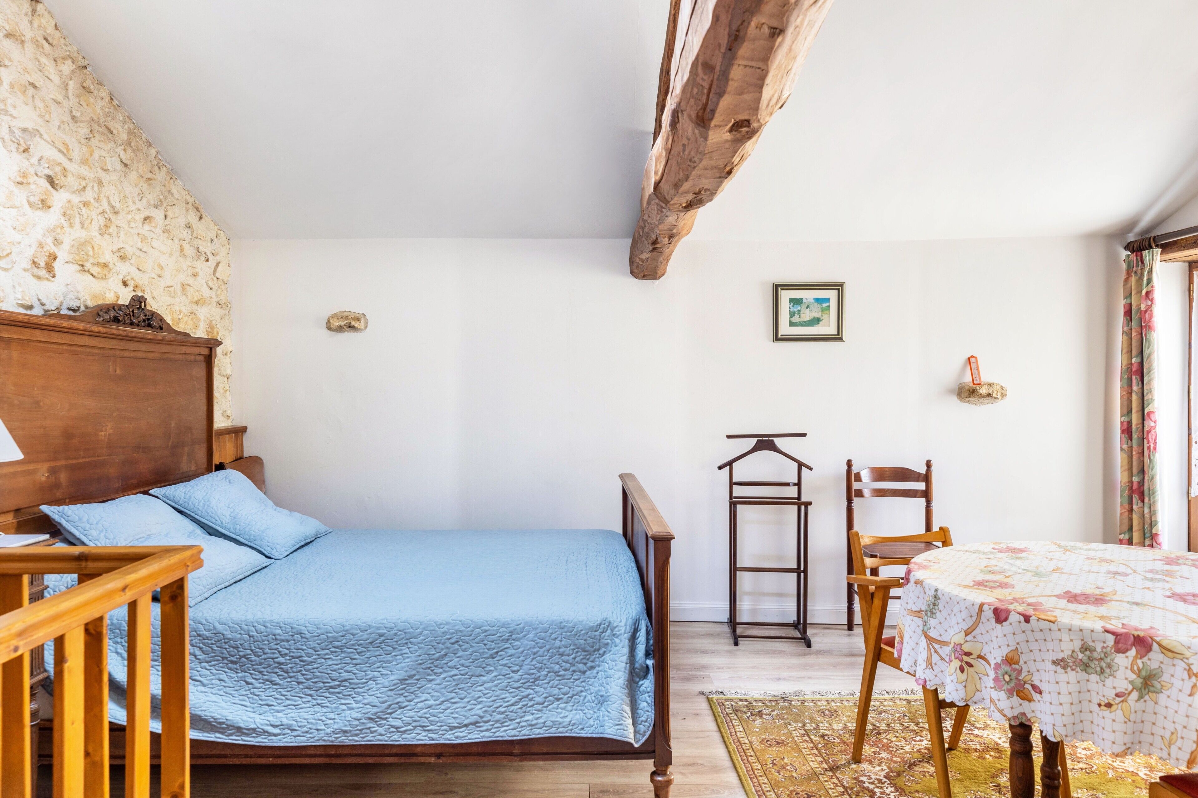 A bedroom with a bed, wooden headboard, a small table with a floral tablecloth, a chair, and a stone wall.