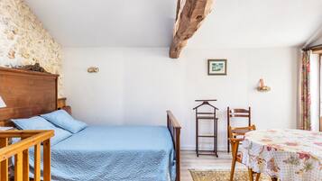 A bedroom with a bed, wooden headboard, a small table with a floral tablecloth, a chair, and a stone wall.