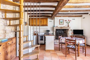 A rustic kitchen with wooden beams, a stone wall, a wooden staircase, a central island, and a dining area with a table and chairs.