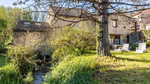 Un edificio de piedra con techo de tejas, un pequeño arroyo y mesas al aire libre.