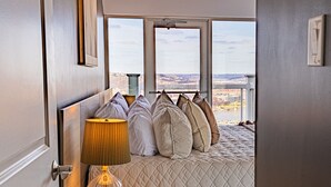 A bedroom with a bed, pillows, a lamp, and a view of a cityscape through large windows. - Luxury Living | Skyline Views | Grandview Retreat (Pittsburgh)