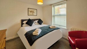 A bedroom with a bed, a red chair, a window with blinds, and a wall-mounted lamp. - Host & Stay | Nile Street Retreat (Norwich)