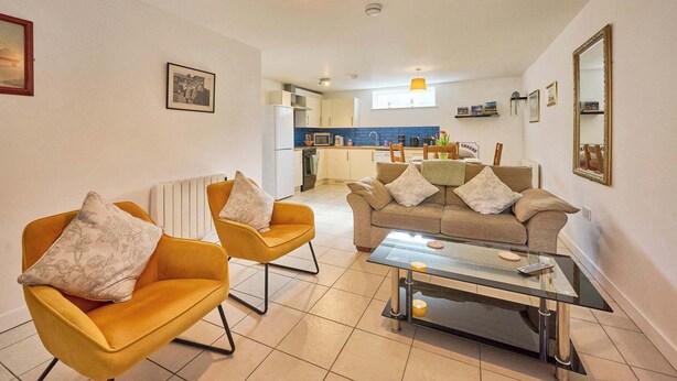 A living room with two yellow armchairs, a sofa, a glass coffee table, and a kitchen in the background. - Host & Stay | Nile Street Retreat (Norwich)