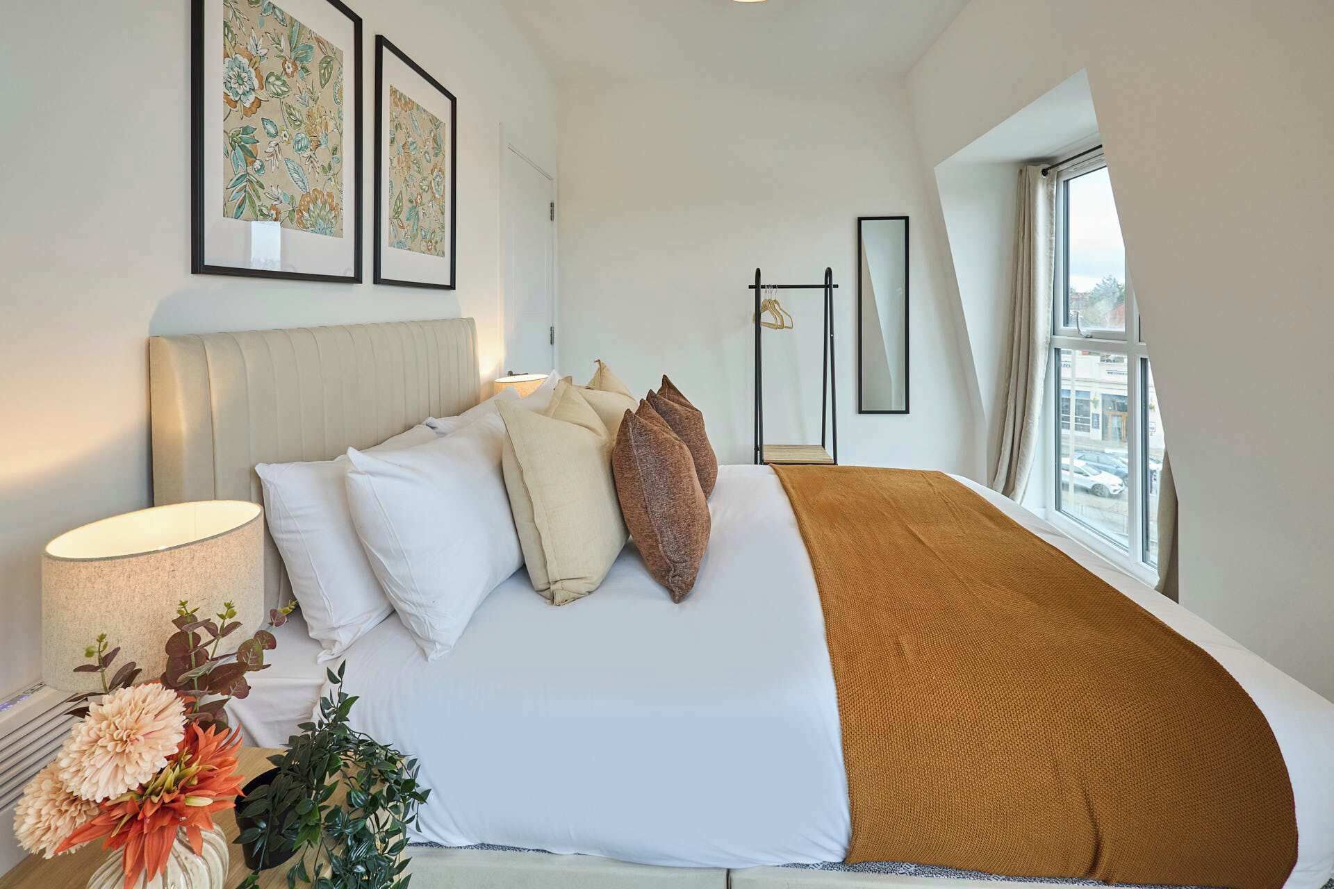 A bedroom with a bed, pillows, a bedside lamp, a window, and framed artwork on the wall.