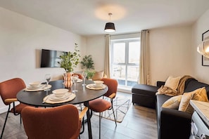 A modern living room with a dining area, a sofa, and a TV mounted on the wall. - Host & Stay | 10 Queens Court (Merseyside)
